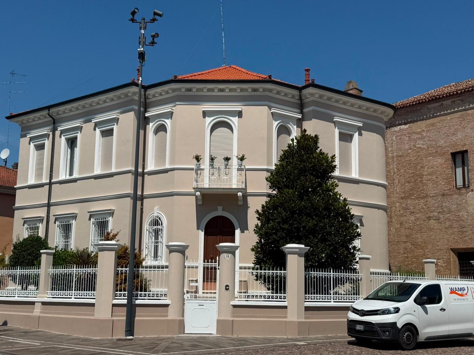 Una casa a due piani color crema con tetto di tegole rosse, recinzione bianca e un furgone parcheggiato sotto un cielo azzurro e limpido.
