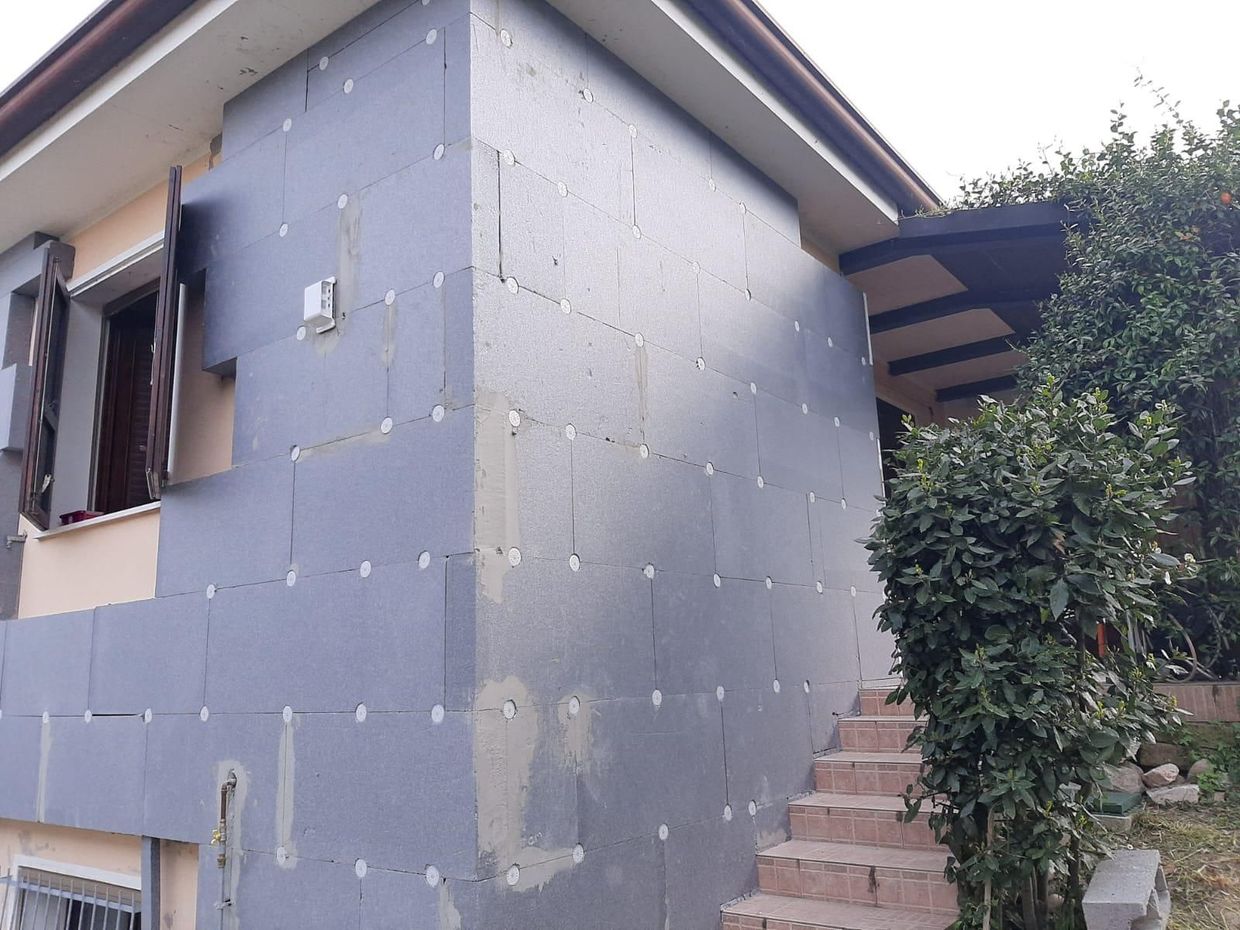 Esterno della casa con pannelli isolanti grigi, finestra aperta e scale accanto a cespugli verdi.