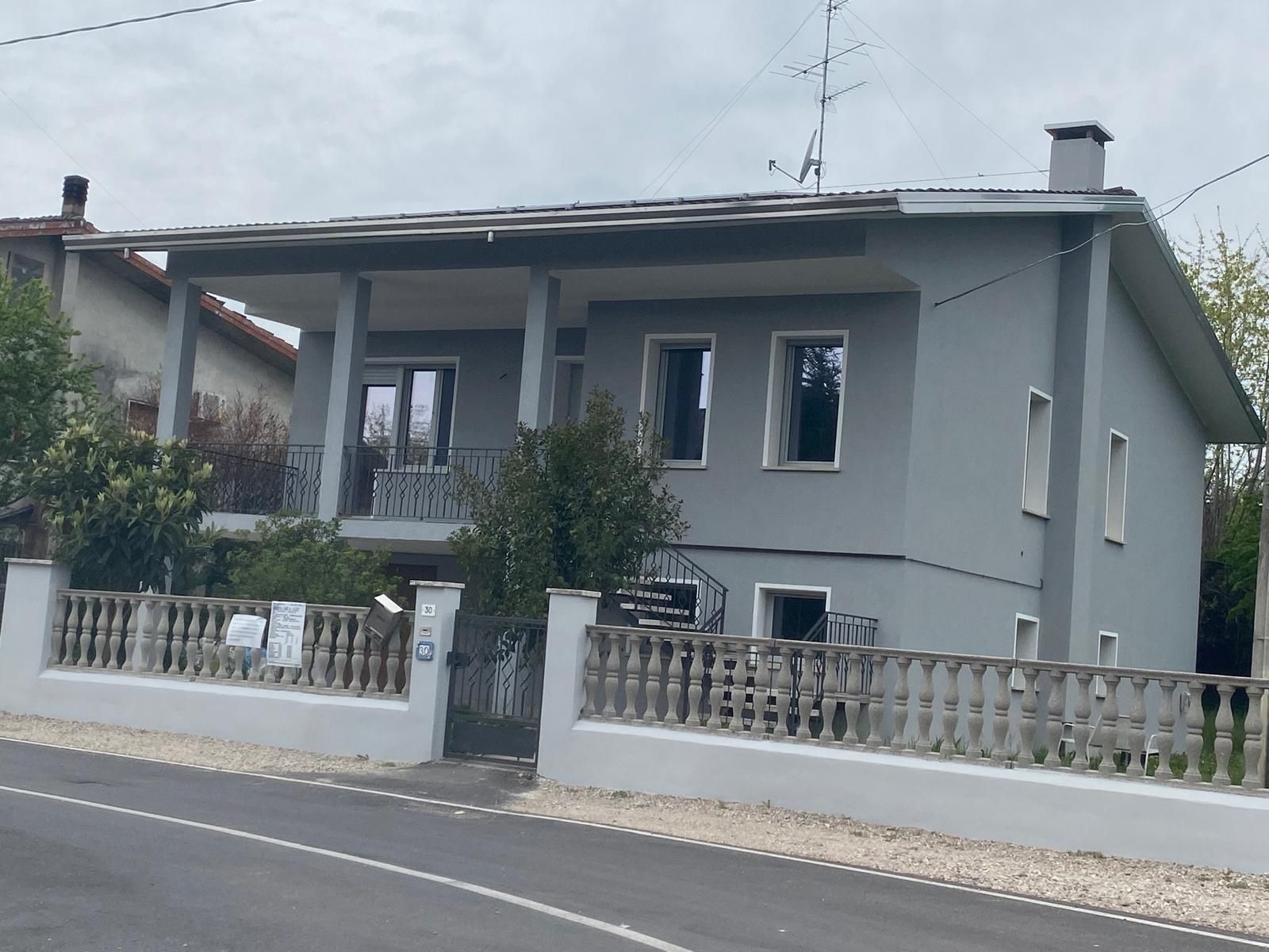 Casa grigia a due piani con recinzione bianca accanto a una strada sotto un cielo nuvoloso