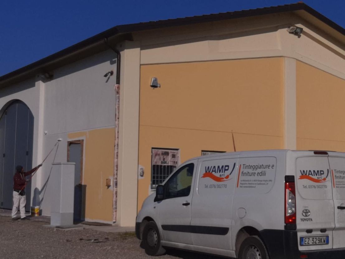 Un operaio dipinge un edificio beige accanto a un furgone bianco in una giornata di sole.