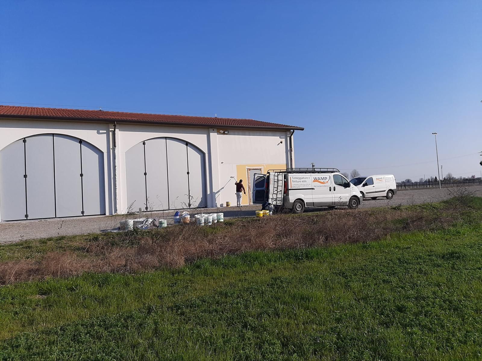Magazzino rurale con grandi portoni grigi e furgoni bianchi parcheggiati accanto a un campo erboso sotto un cielo azzurro.