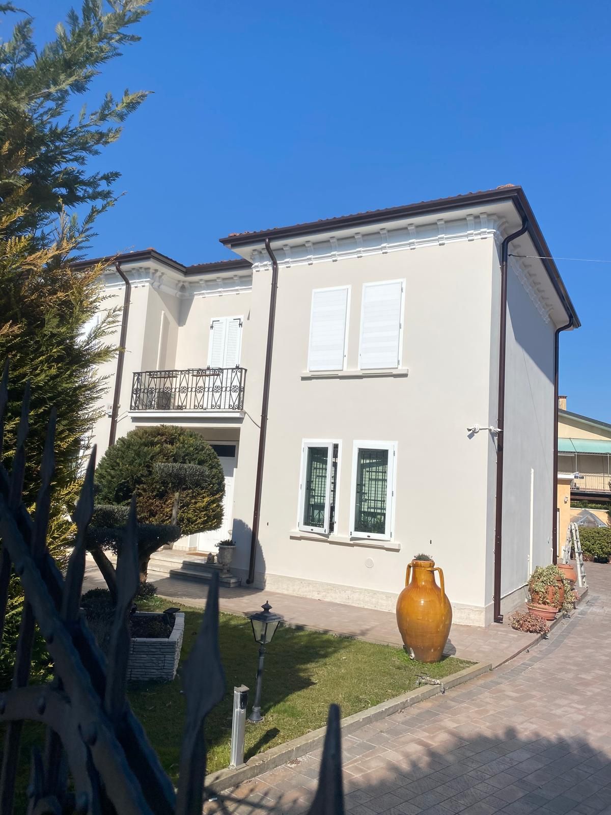 Casa color crema a due piani con balcone, giardino e vialetto d'accesso sotto un cielo azzurro brillante