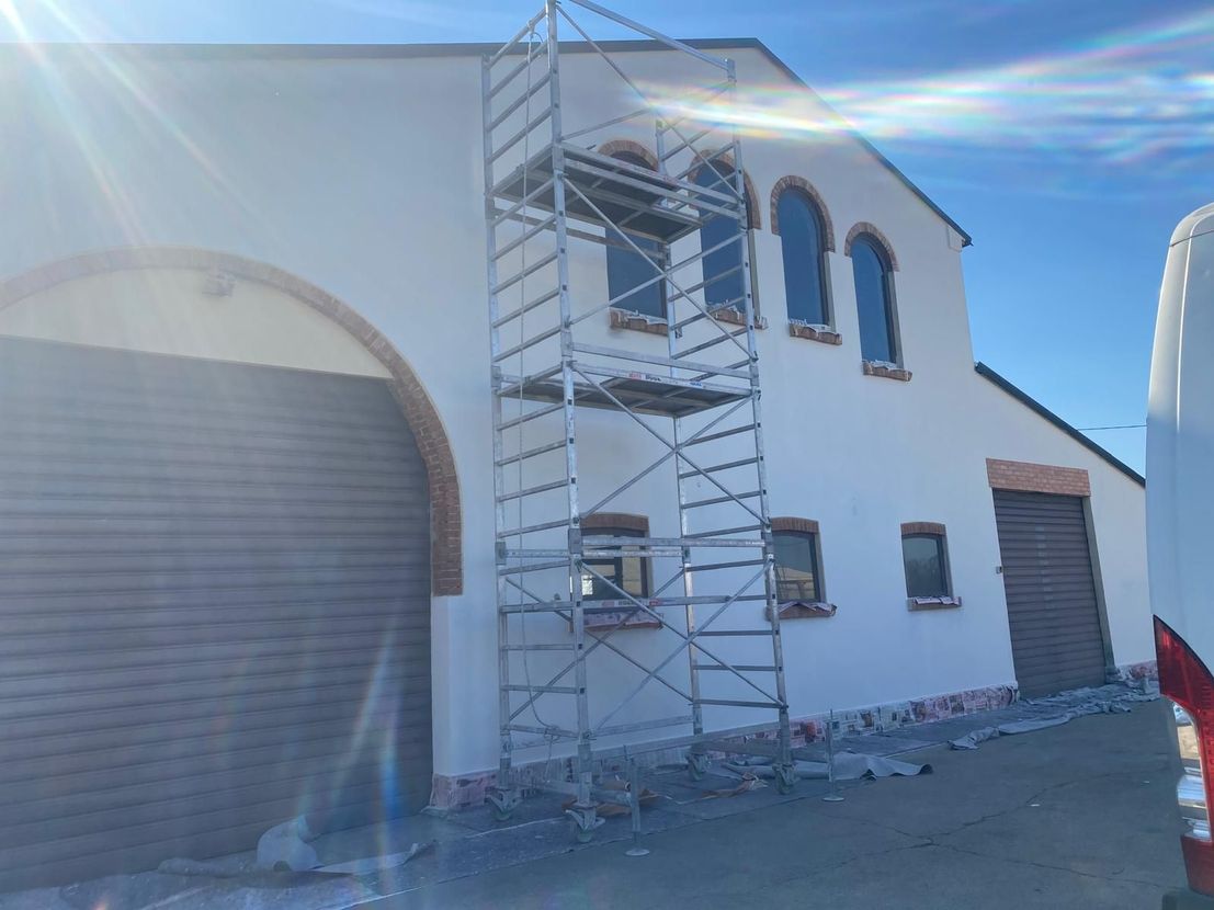 Edificio bianco con impalcature accanto a un grande portone da garage, illuminato da un sole splendente.