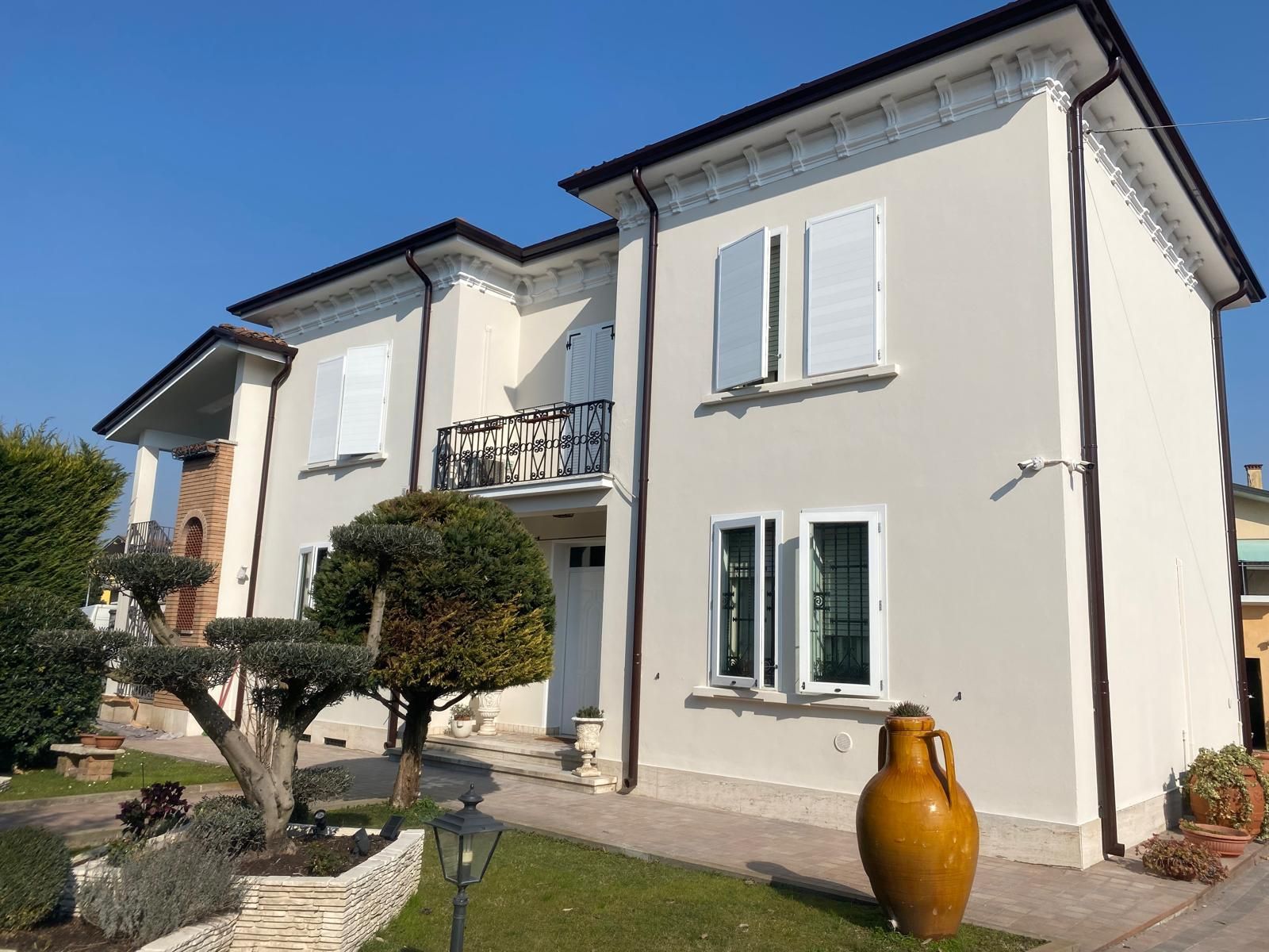 Casa bianca a due piani con un piccolo balcone, alberi potati e una grande urna gialla nel giardino anteriore.