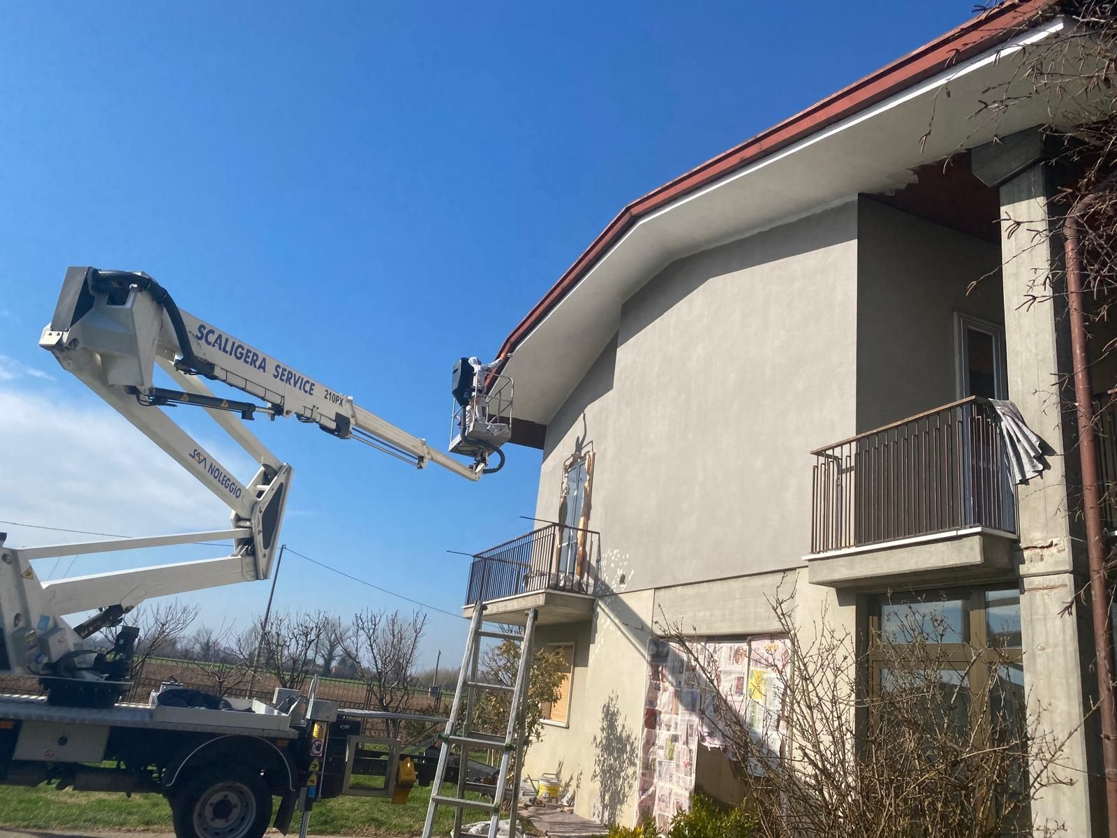 Un operaio, all'interno di una piattaforma elevatrice, ripara la cornice del tetto di una casa beige in una giornata di sole.