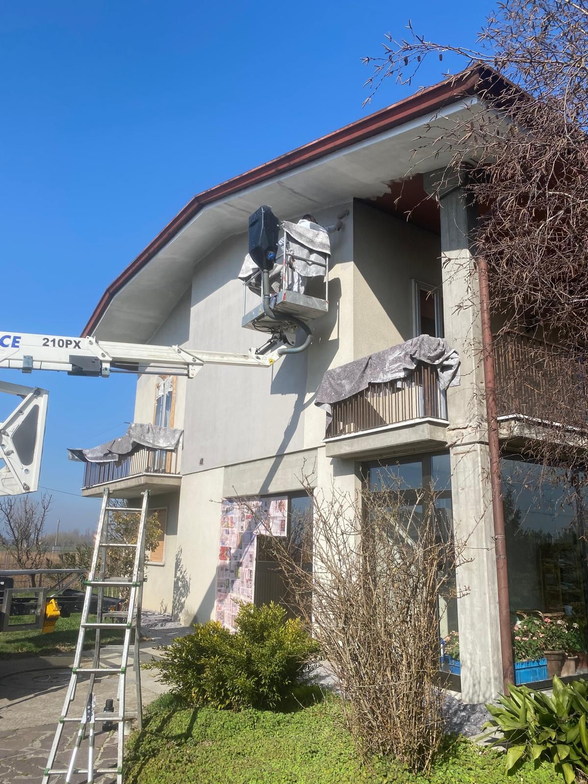 Due operai su una scala dipingono l'esterno di una casa, con una piattaforma aerea nelle vicinanze, sotto un cielo azzurro.
