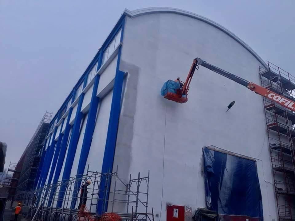 Edificio industriale con struttura in acciaio blu e operai su una piattaforma elevatrice accanto a una parete bianca curva.