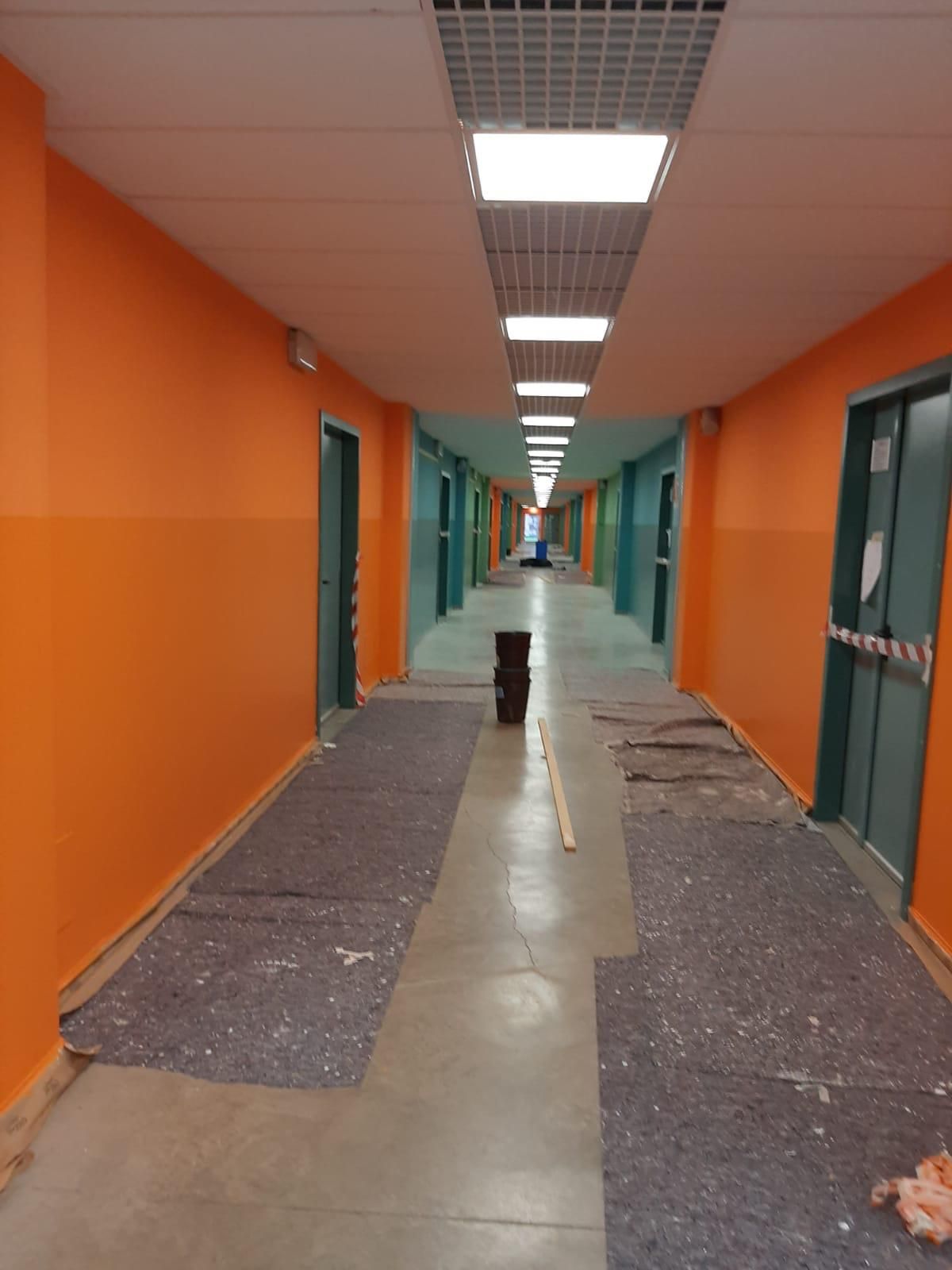 Corridoio arancione con porte color verde acqua, luci a soffitto e rivestimenti del pavimento strappati in un lungo corridoio.
