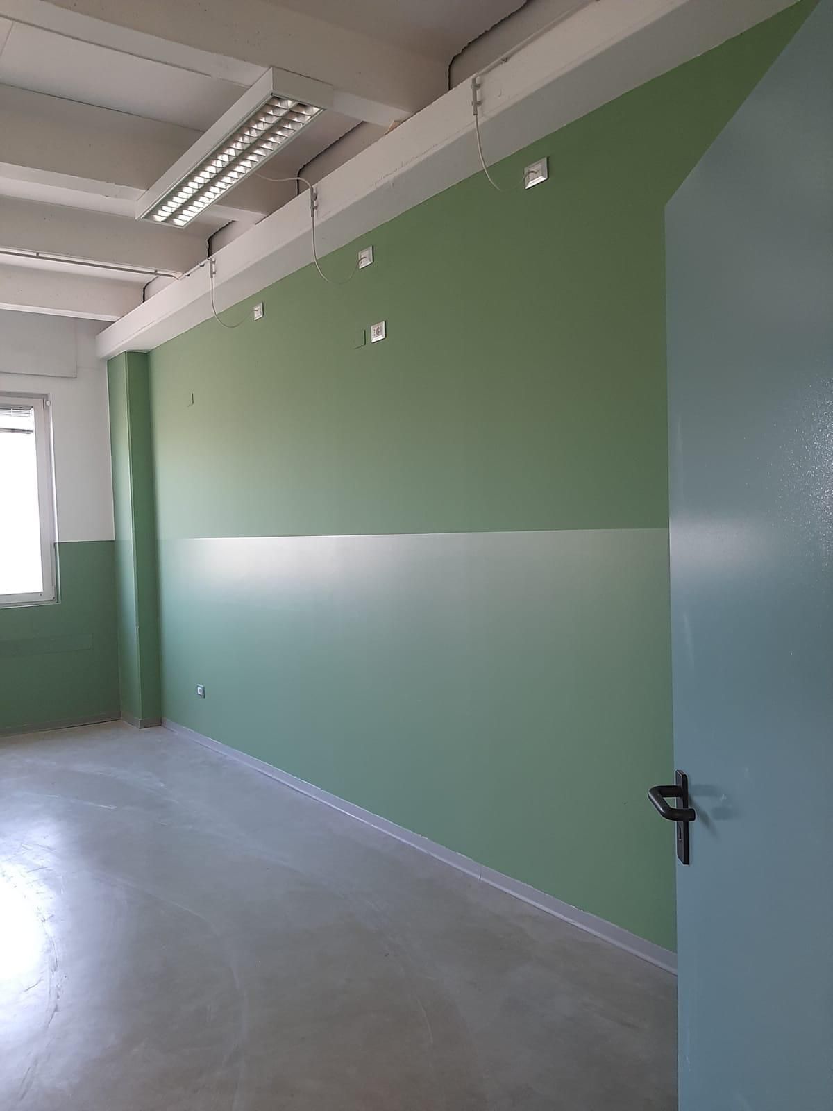 Corridoio verde vuoto con pavimento bianco, luci a soffitto e una porta aperta sulla destra.