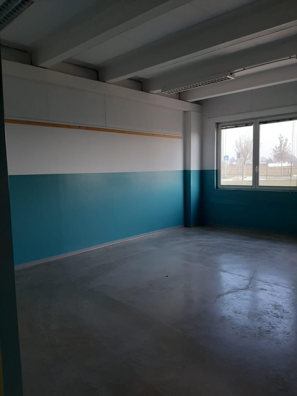 Stanza vuota con pareti color verde acqua e bianche, pavimento in cemento, travi a vista sul soffitto e una finestra sulla destra.