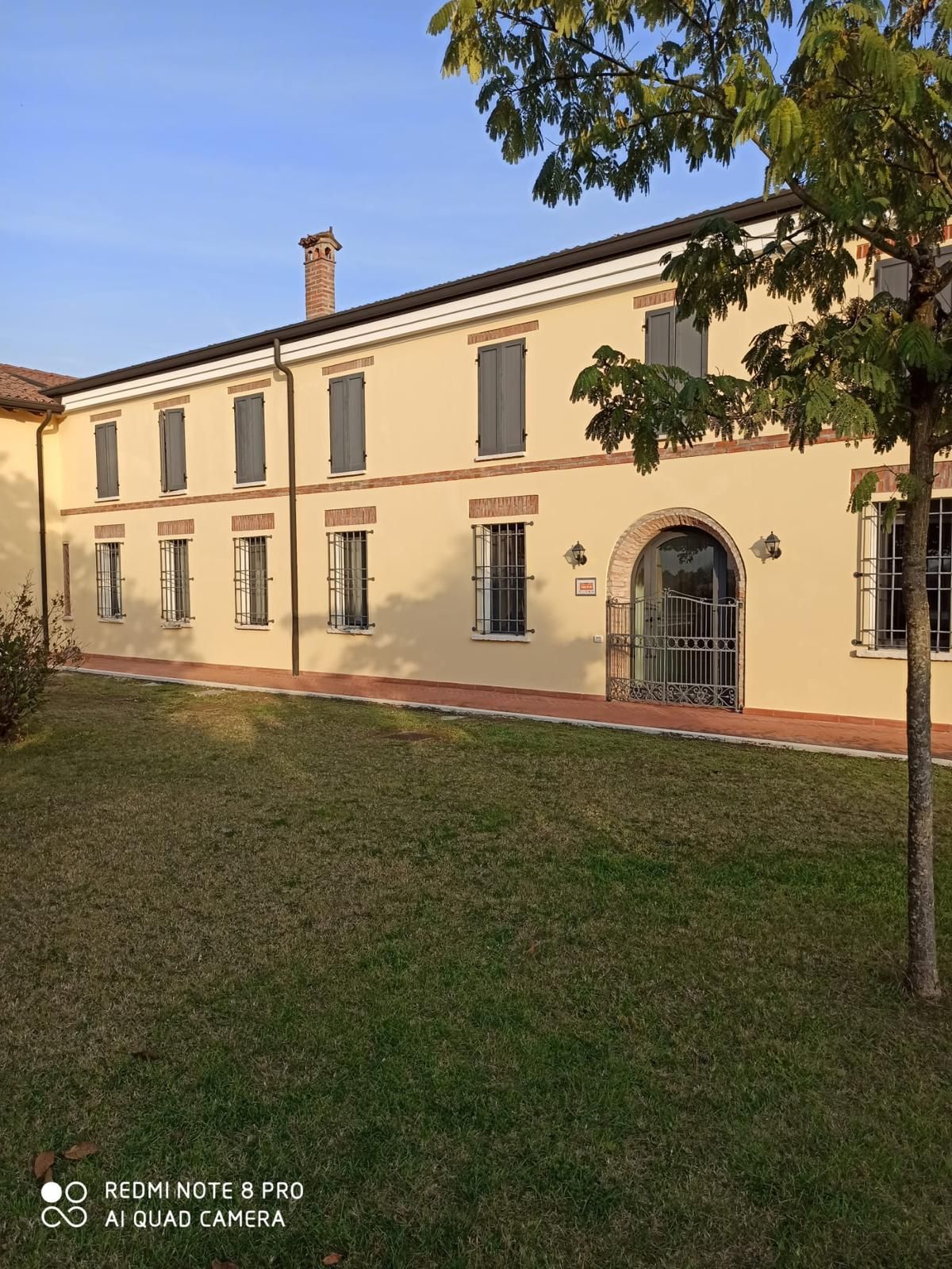 Edificio color crema a due piani con finestre con persiane e cancello ad arco, accanto a un prato e un albero sotto un cielo azzurro.