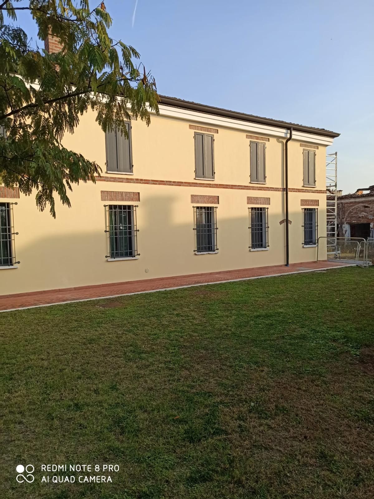 Edificio color crema a due piani con persiane blu accanto a un prato al tramonto.