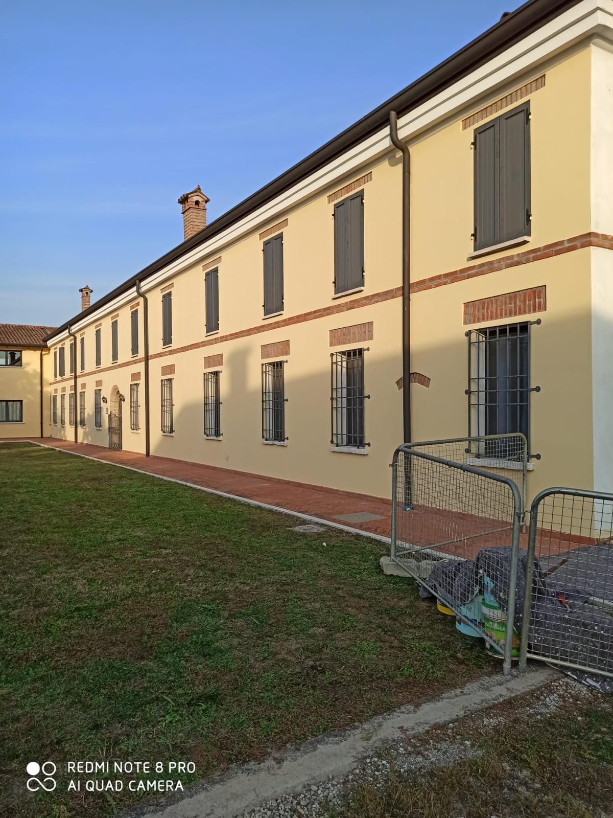 Edificio giallo illuminato dal sole con finestre con persiane e un cortile erboso, visto da un'angolazione