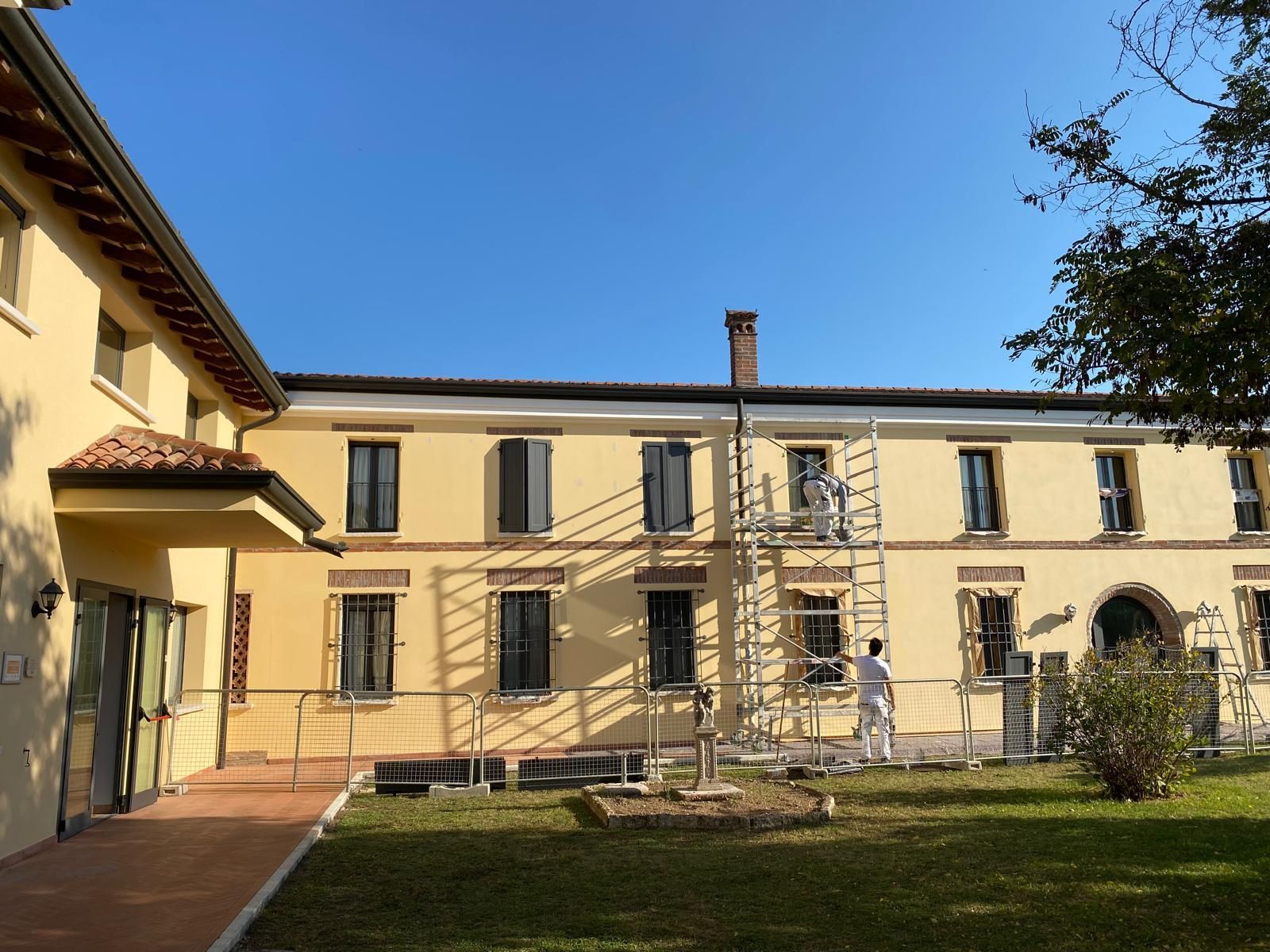 Cortile soleggiato di un edificio giallo con finestre ad arco e alberi sotto un cielo azzurro limpido.