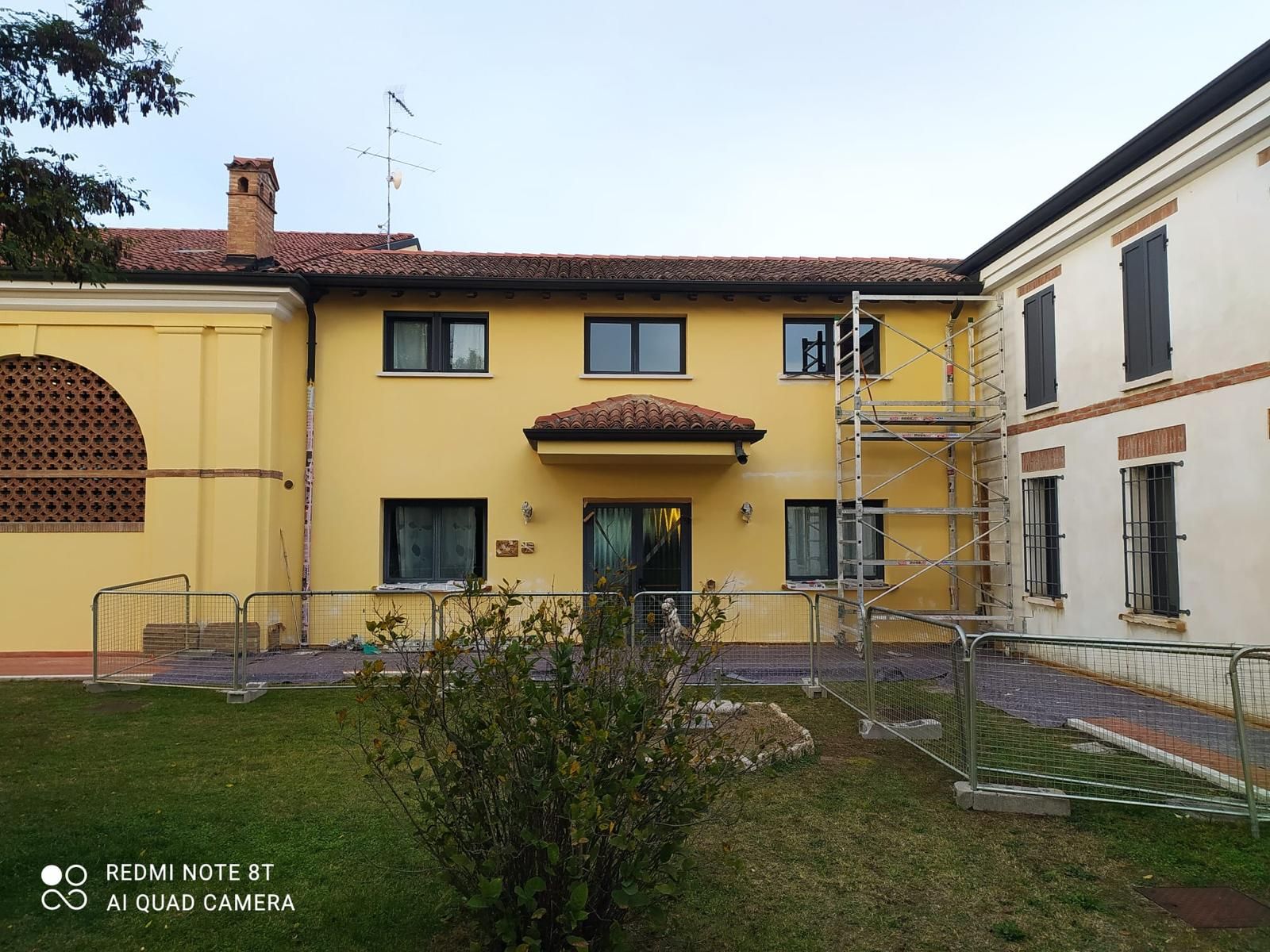 Casa gialla a due piani con tetto di tegole e piccolo cortile con giardino, fiancheggiata da edifici adiacenti e impalcature.