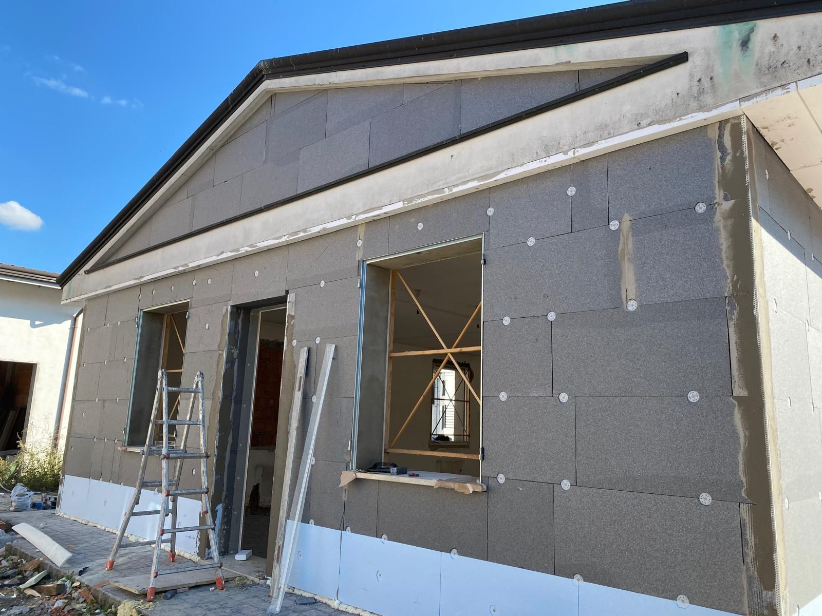 Esterno di una casa parzialmente ultimato con rivestimento grigio, infissi di porte e finestre ancora aperti e impalcature da cantiere.