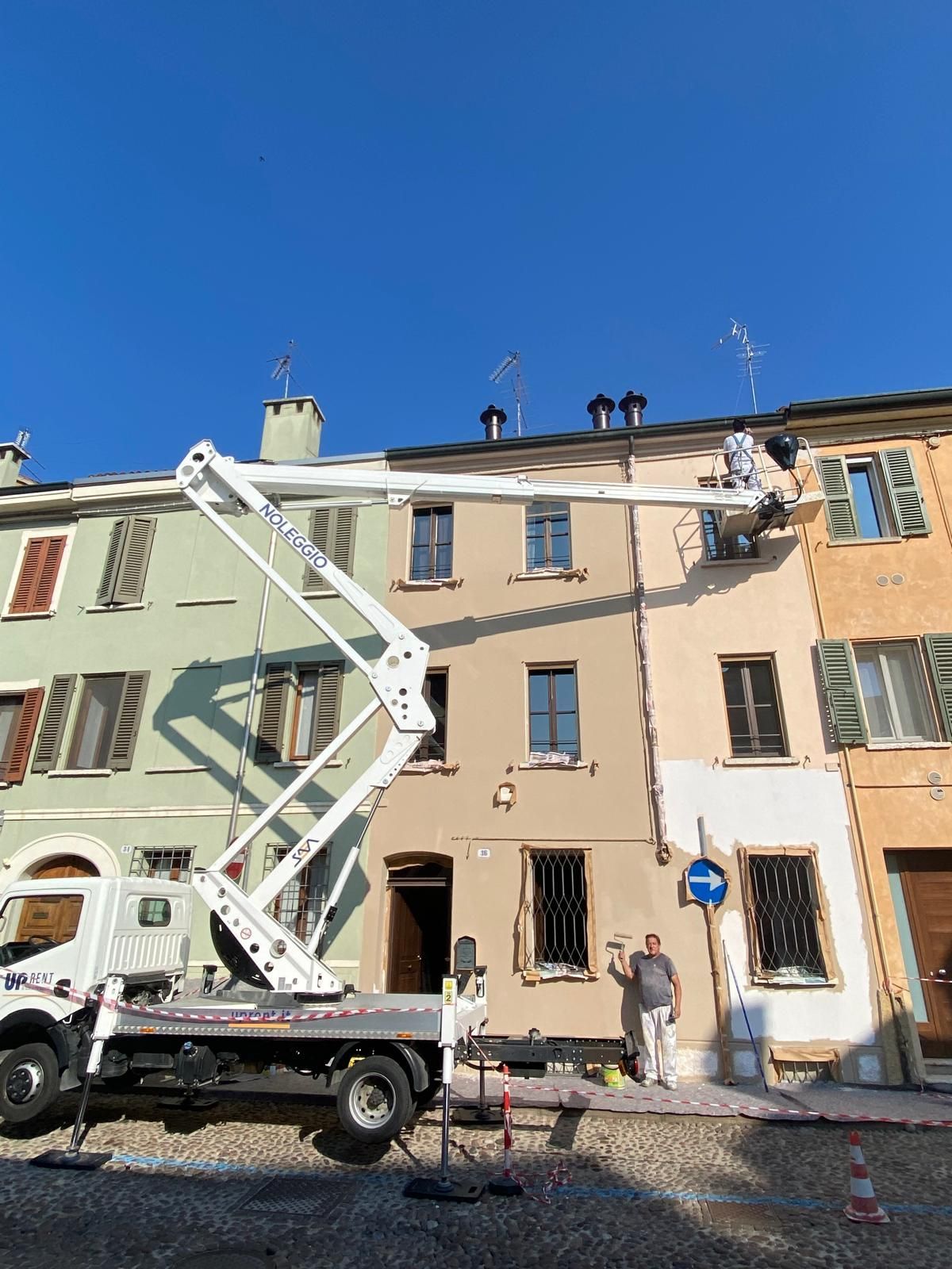 Operai su una piattaforma elevatrice riparano il tetto di un edificio che ospita un pesco, sotto un cielo azzurro e limpido, in una piazza cittadina.