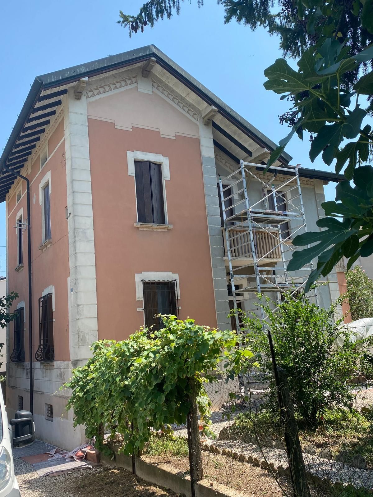 Casa rosa in costruzione con impalcature accanto a un sentiero del giardino e all'ombra di un albero.