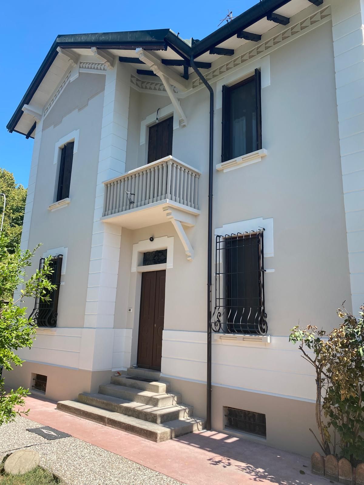 Casa bianca a due piani con un piccolo balcone, tetto scuro e scalini d'ingresso esposti alla luce del sole.
