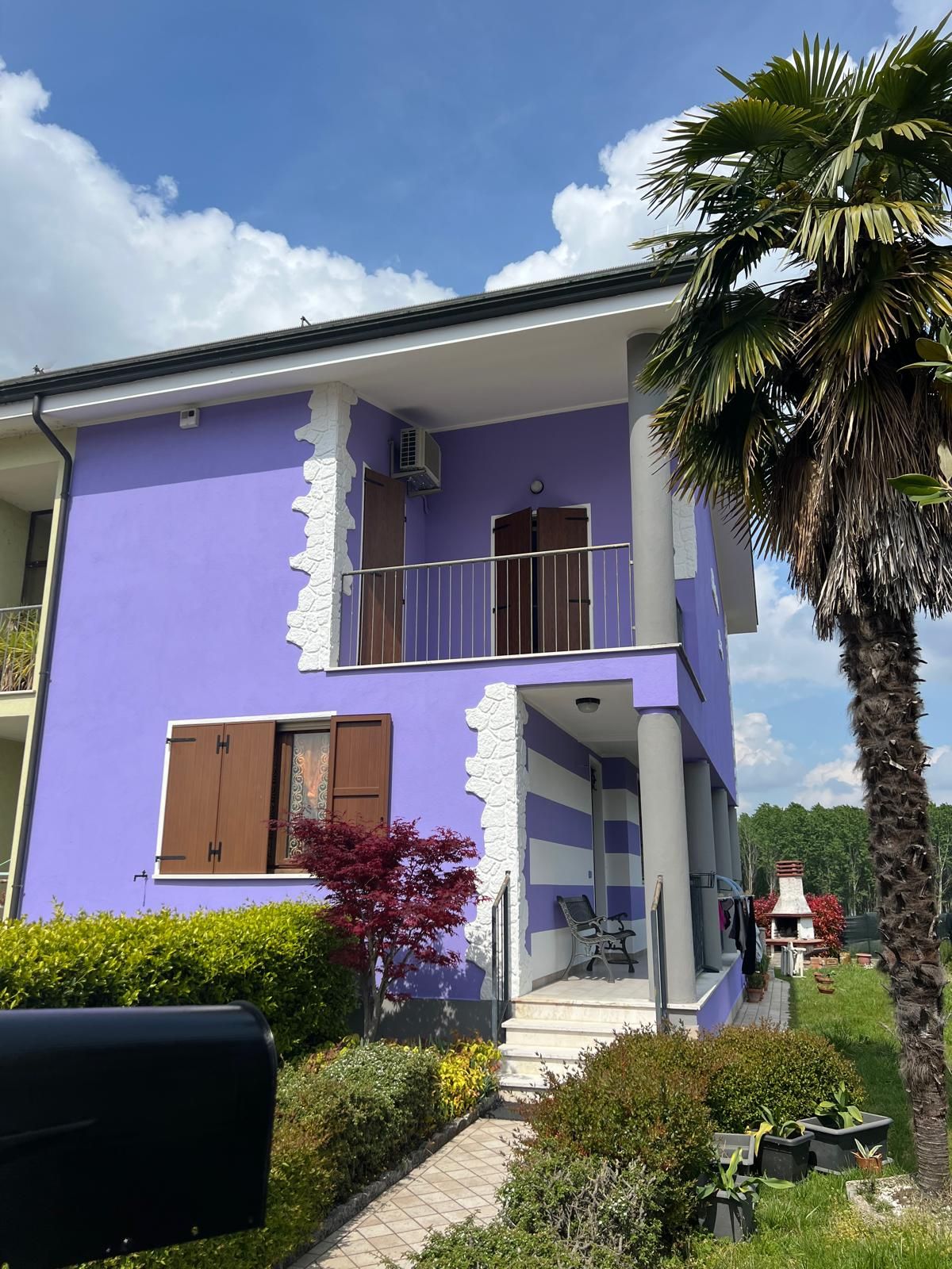 Casa viola a due piani con balcone, persiane marroni e una palma davanti, sotto un cielo azzurro.
