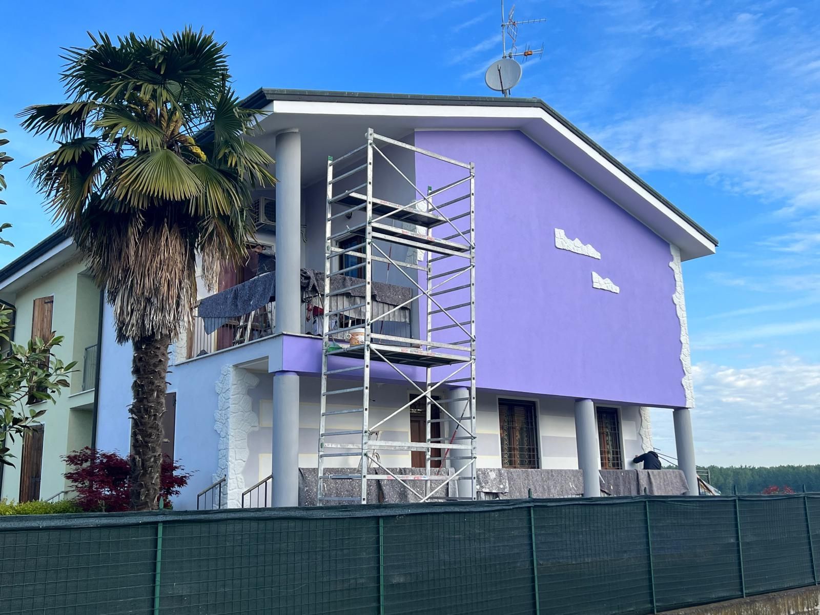 Edificio viola con impalcature all'esterno, accanto a una palma sotto un cielo azzurro.