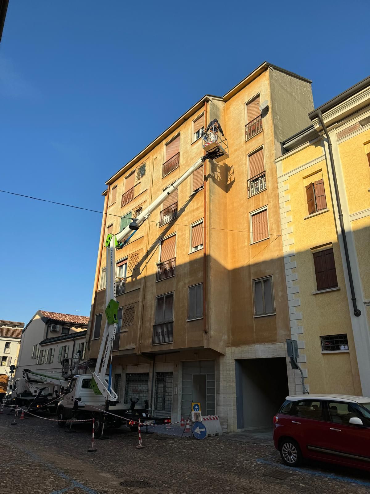 Un palazzo giallo illuminato dal sole su una strada cittadina, con auto parcheggiate e un camion di servizio che solleva attrezzature sottostante.