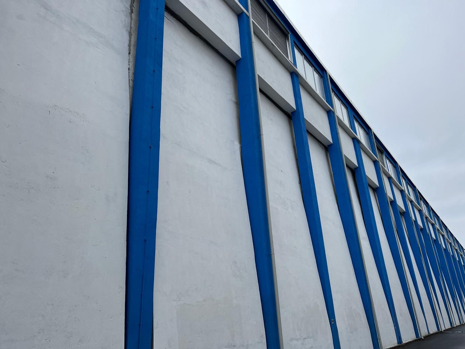 Una recinzione metallica blu e bianca si perde lungo il muro di un edificio sotto un cielo nuvoloso.