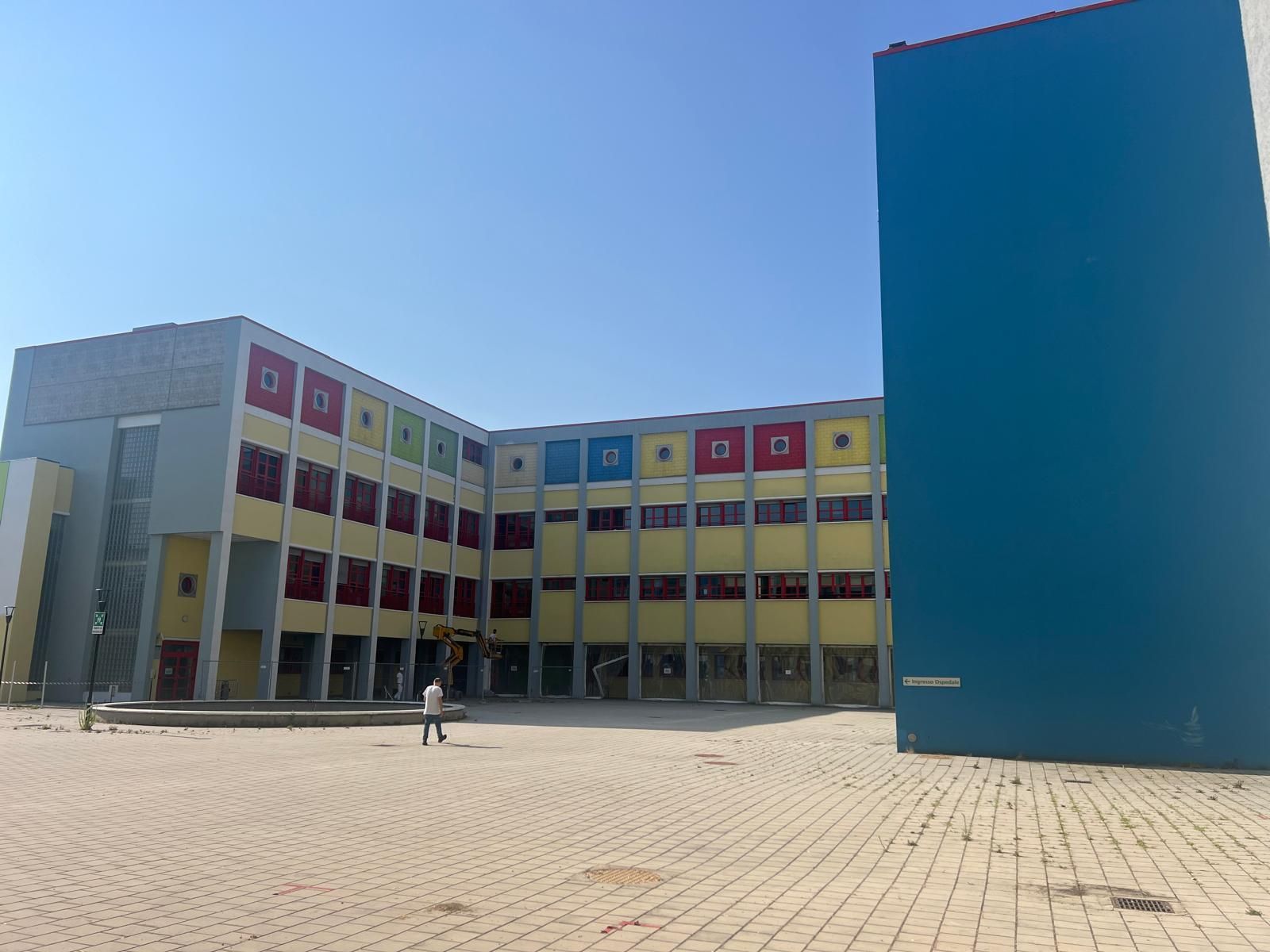 Cortile scolastico moderno con un edificio giallo e rosso e un alto muro blu sotto un cielo limpido