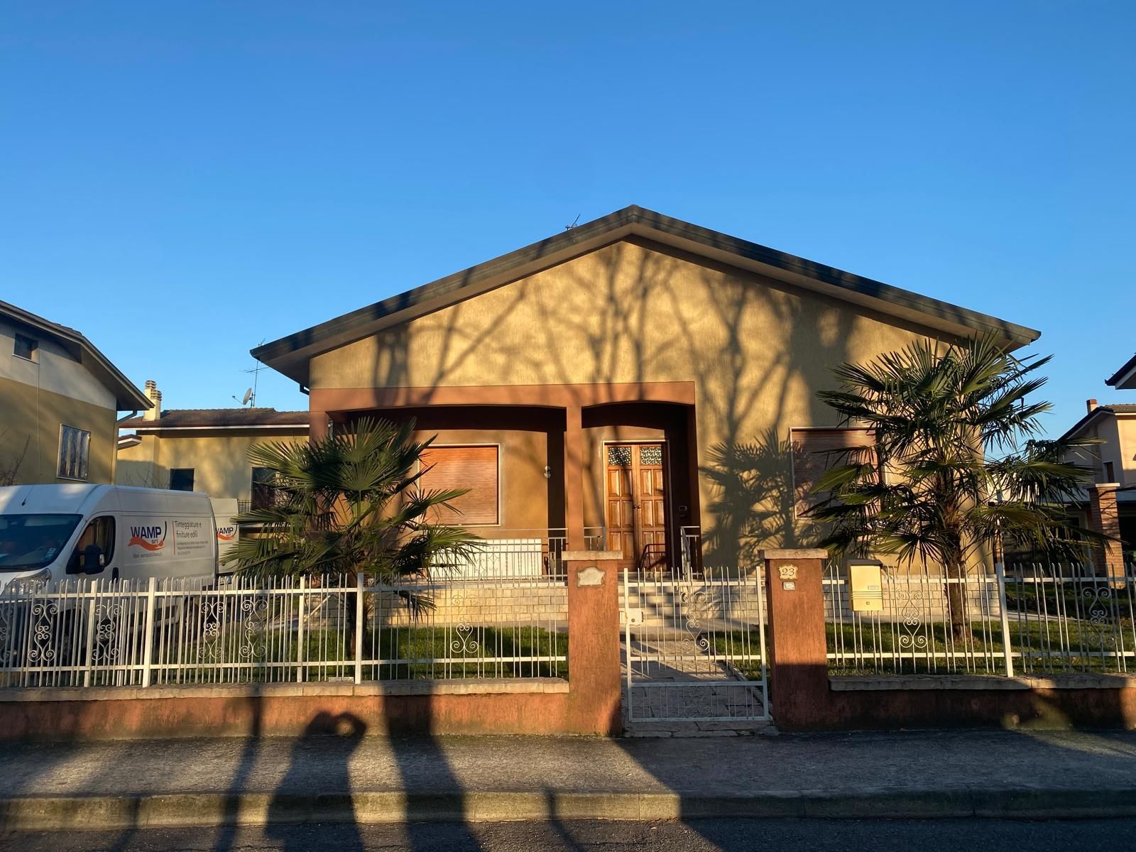 Casa a un solo piano color beige con cancello d'ingresso e ombre di alberi spogli, vista dalla strada.