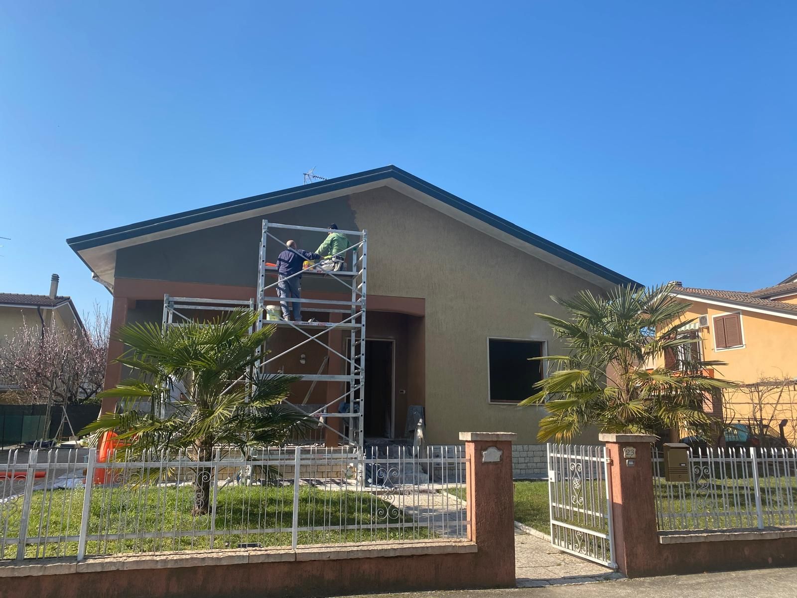 Casa in costruzione con impalcature davanti, cortile recintato e cielo azzurro limpido.