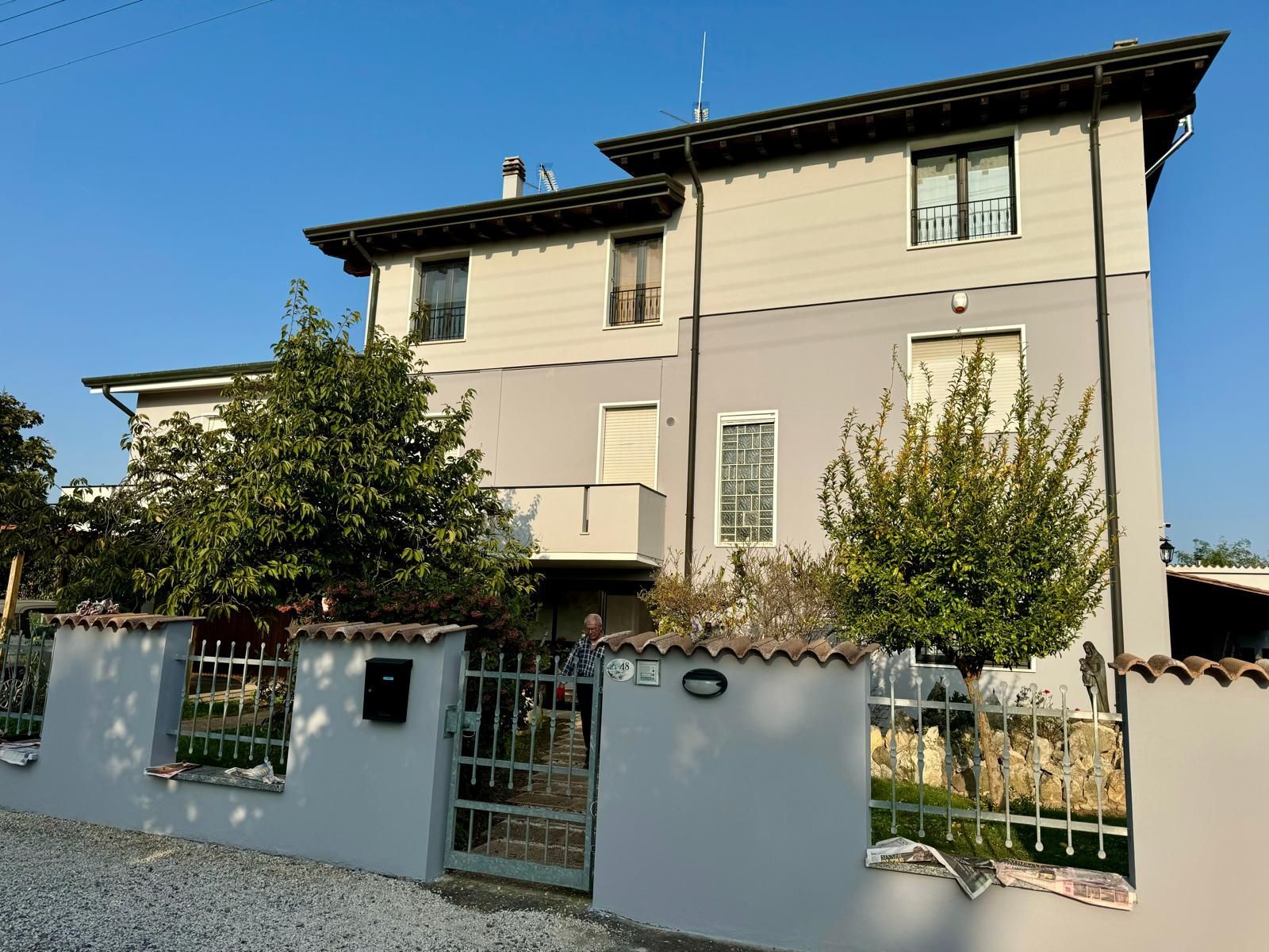 Casa beige a due piani con recinzione bianca, cancello e alberi di fronte, sotto un cielo azzurro limpido.