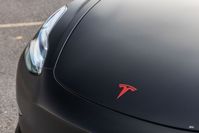 Close-up of a matte black Tesla car hood with a red Tesla emblem.