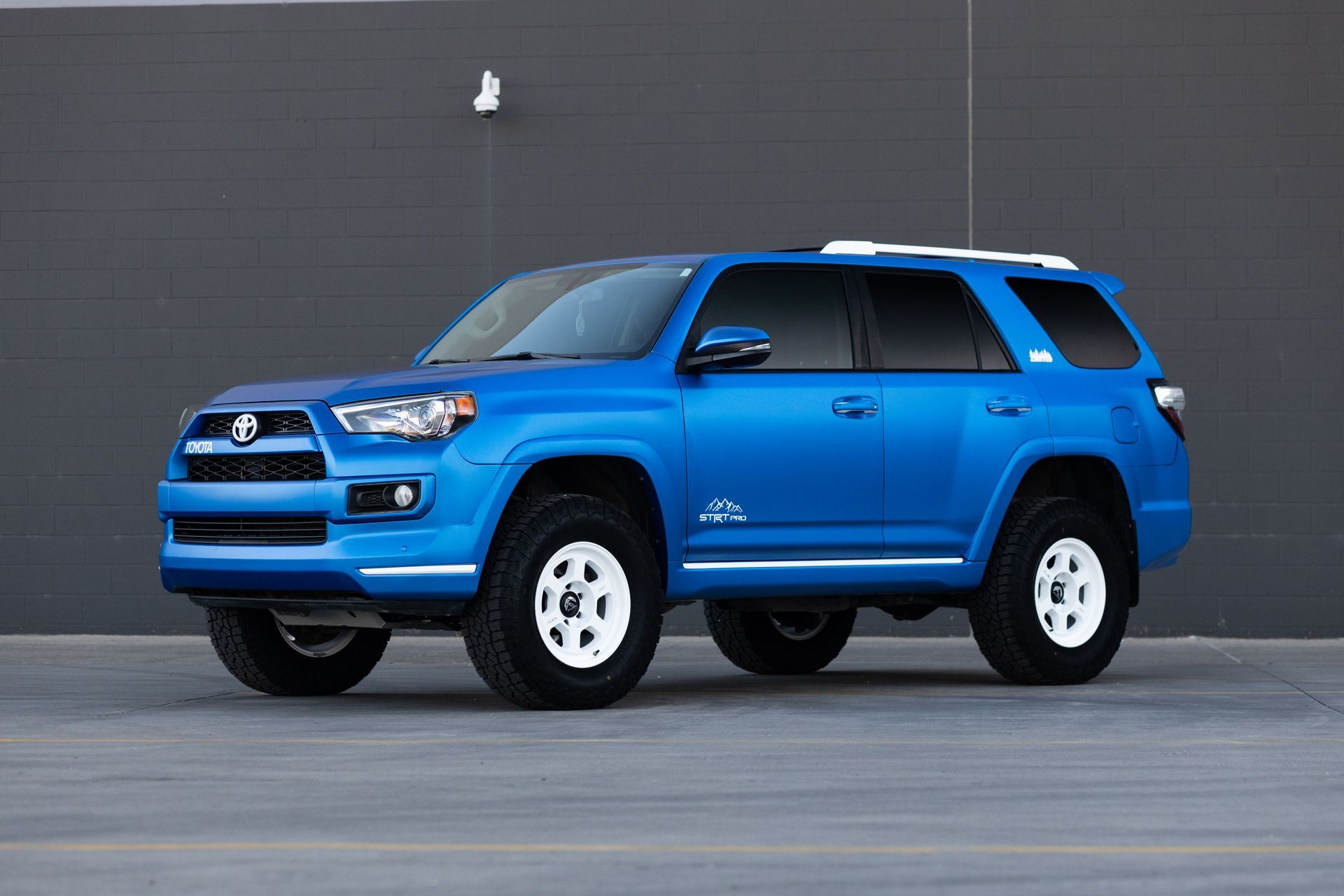 Blue Toyota 4Runner SUV with white wheels, parked in front of a gray wall.