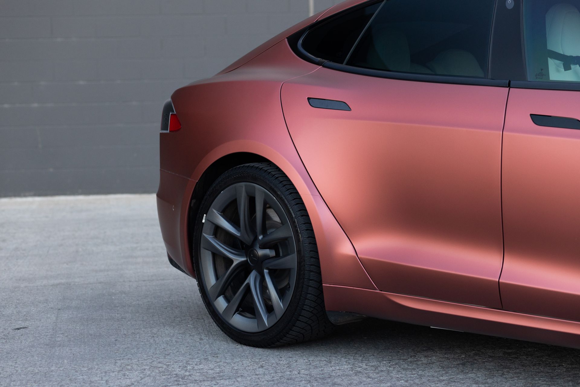 Rear quarter of a bronze Tesla sedan with black wheels parked on concrete.