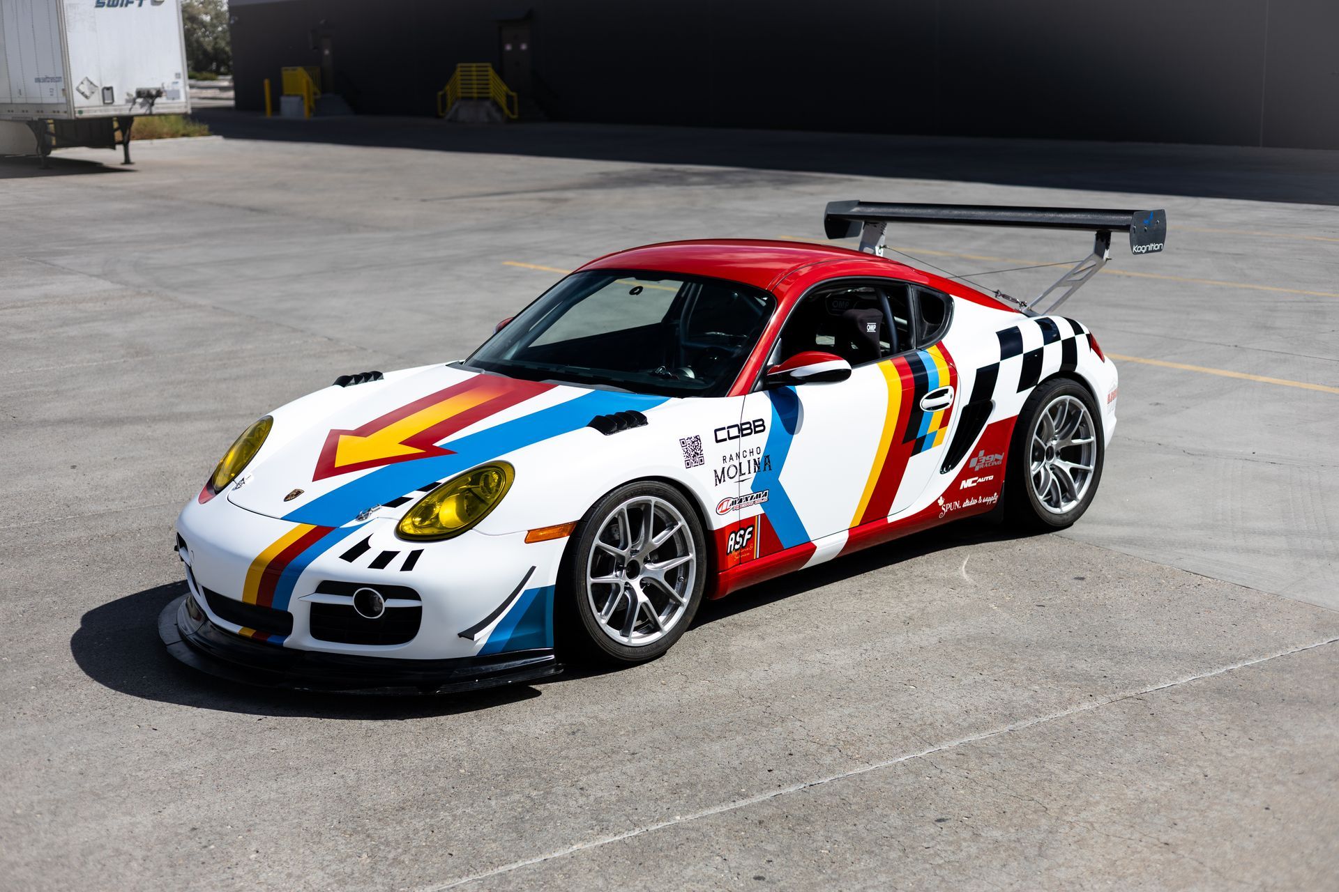 Race car, white Porsche Cayman with red, blue, and yellow racing livery, black spoiler, on asphalt.