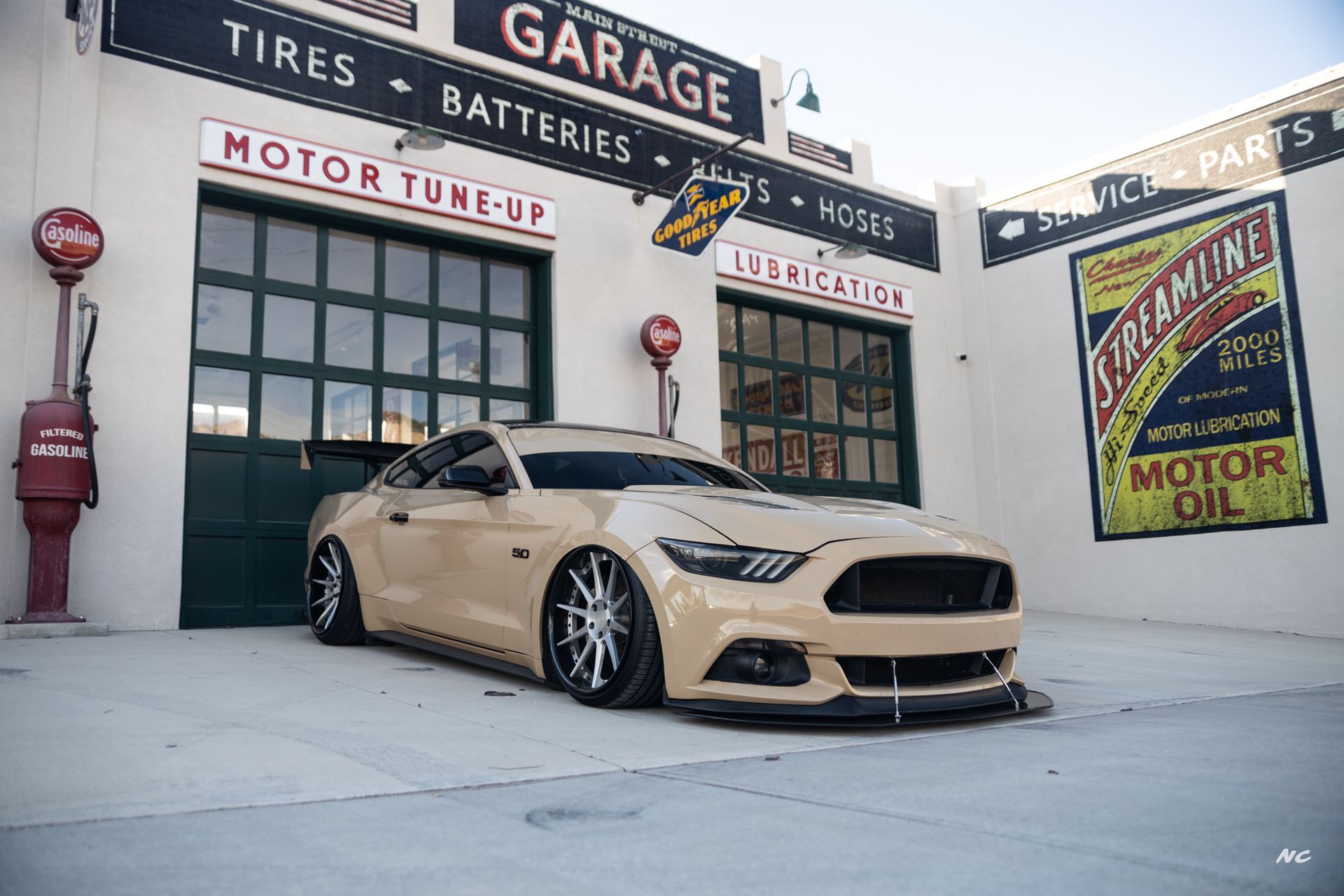Tan Mustang in front of an old garage with signs for tires, batteries, and oil.