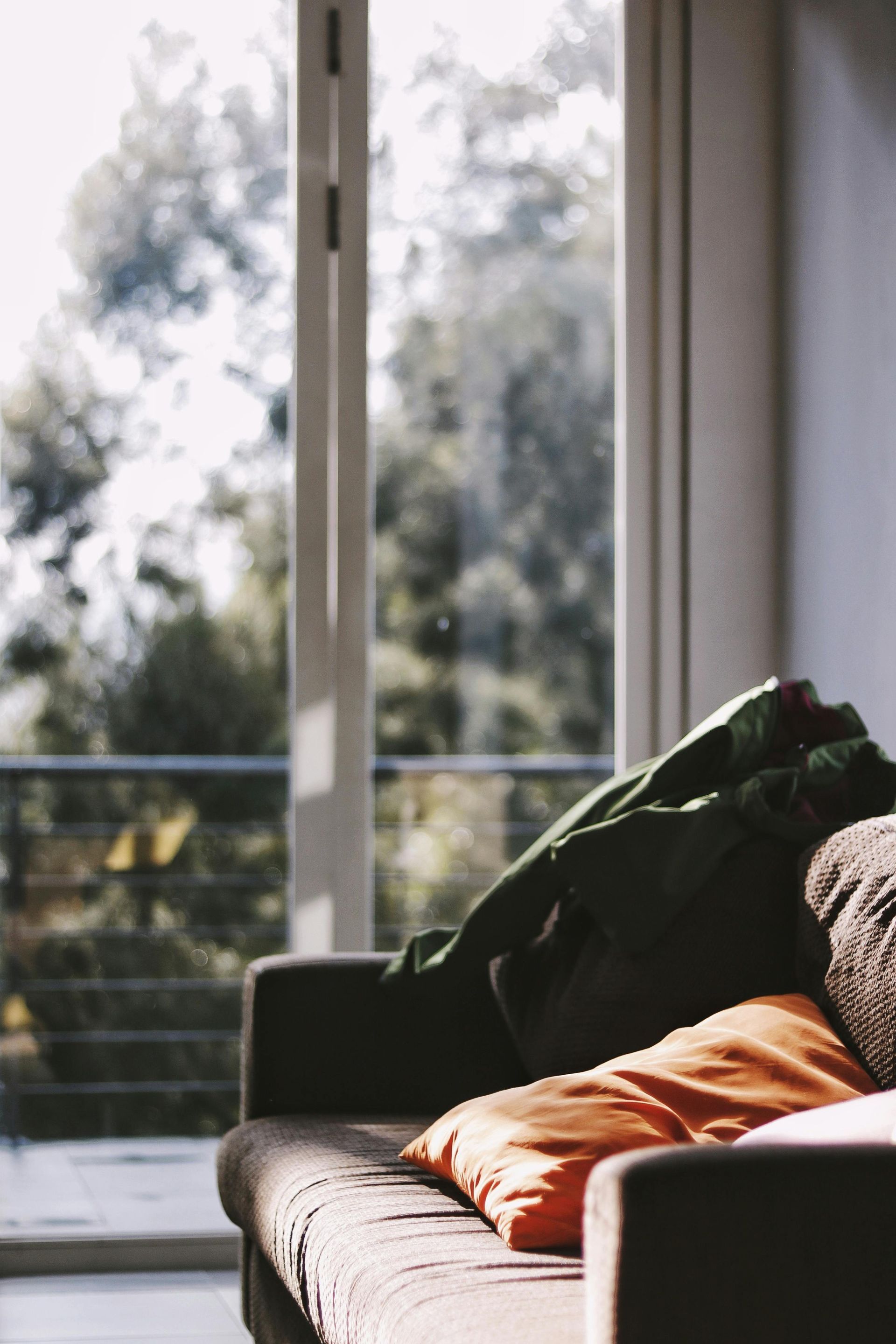 Couch with orange and green pillows near a window, overlooking a blurred green tree backdrop.