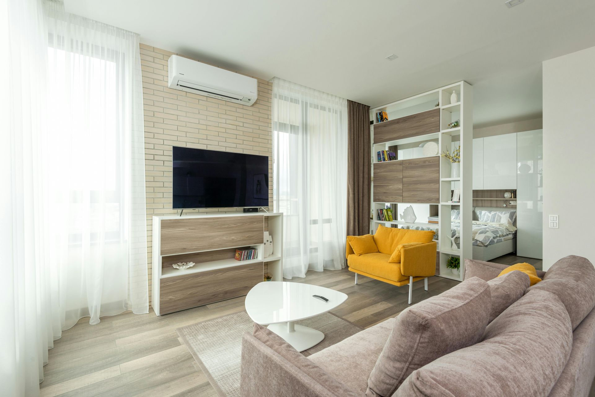 Bright living room with TV, white shelves, yellow chair, and brown sofa near sheer curtains