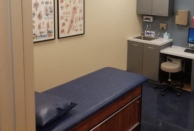 A small medical exam room with a dark blue examination table, a sink, cabinetry, a computer desk, and anatomy wall charts.