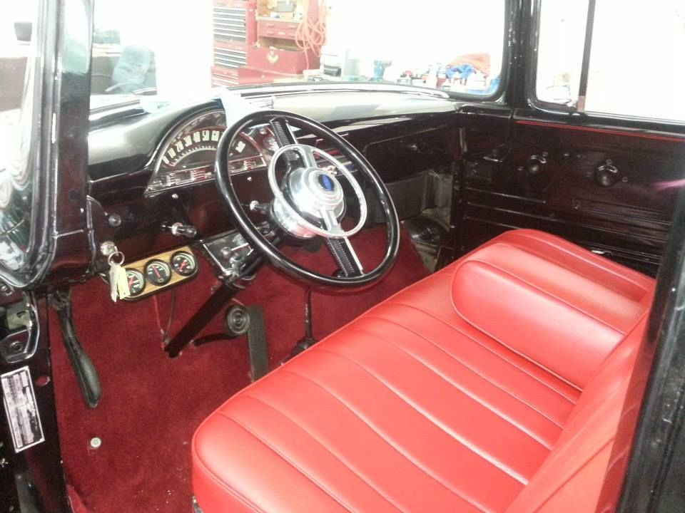 The interior of a classic truck featuring a red bench seat, black steering wheel, and maroon carpeting.