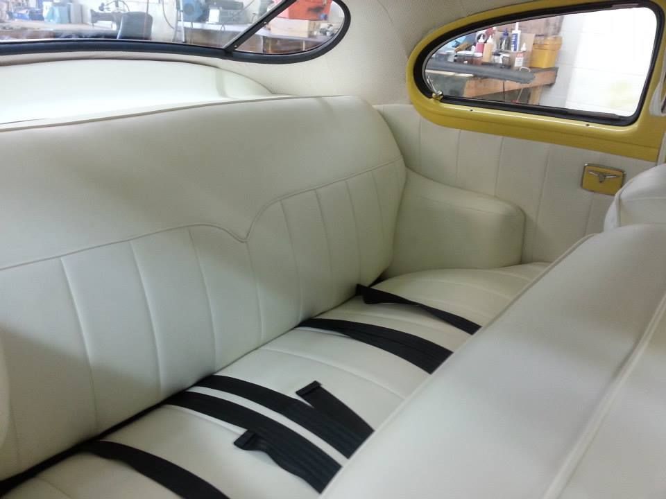 The back seat of a car features clean, white tufted upholstery and black seatbelts, with a yellow-trimmed side window.