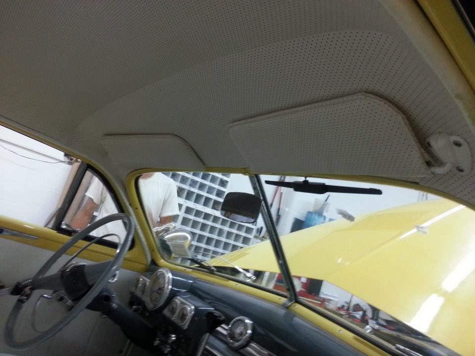 Interior view of a classic yellow car, showing the dashboard, steering wheel, sun visors, and a perforated headliner.