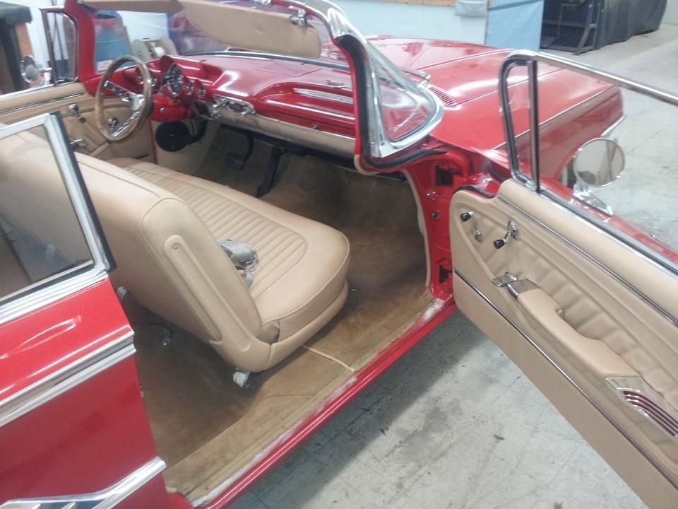 Interior of a classic bright red convertible car with tan bench seating, parked in a garage with the driver door open.