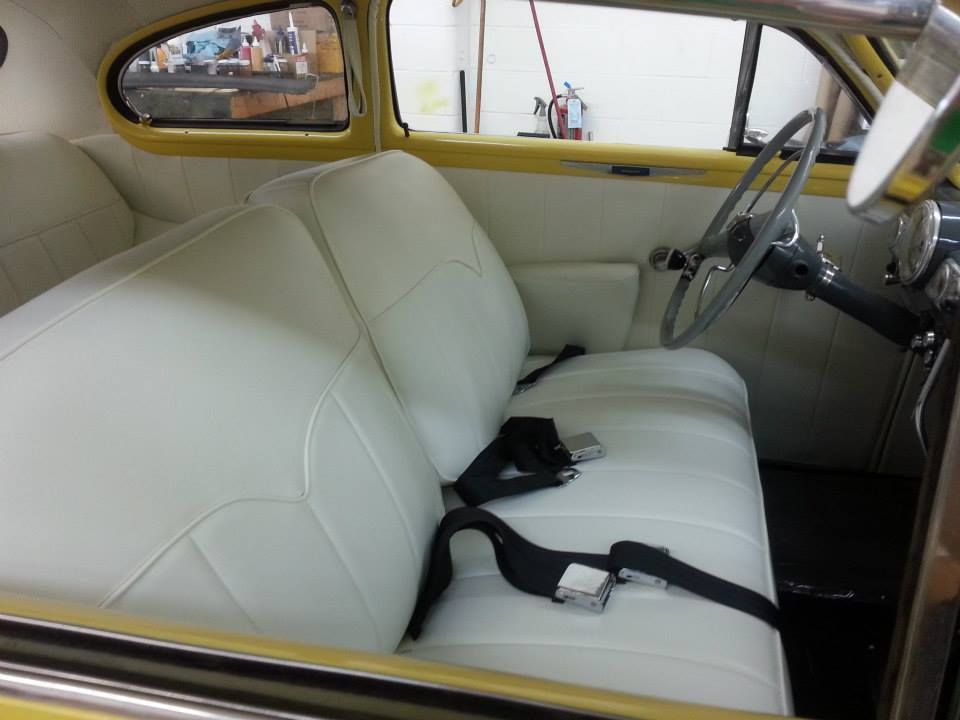 Interior of a classic car with white bench seats, black seatbelts, a large steering wheel, and yellow door trim.