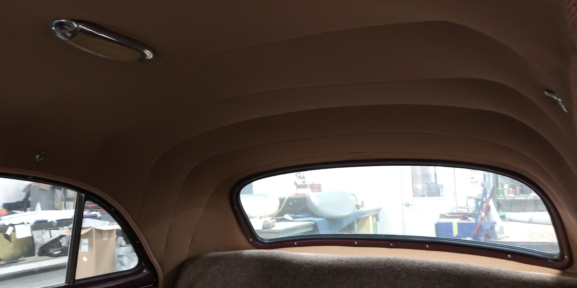Interior view of a vintage car showing a tan fabric headliner, a chrome dome light, and the rear window.