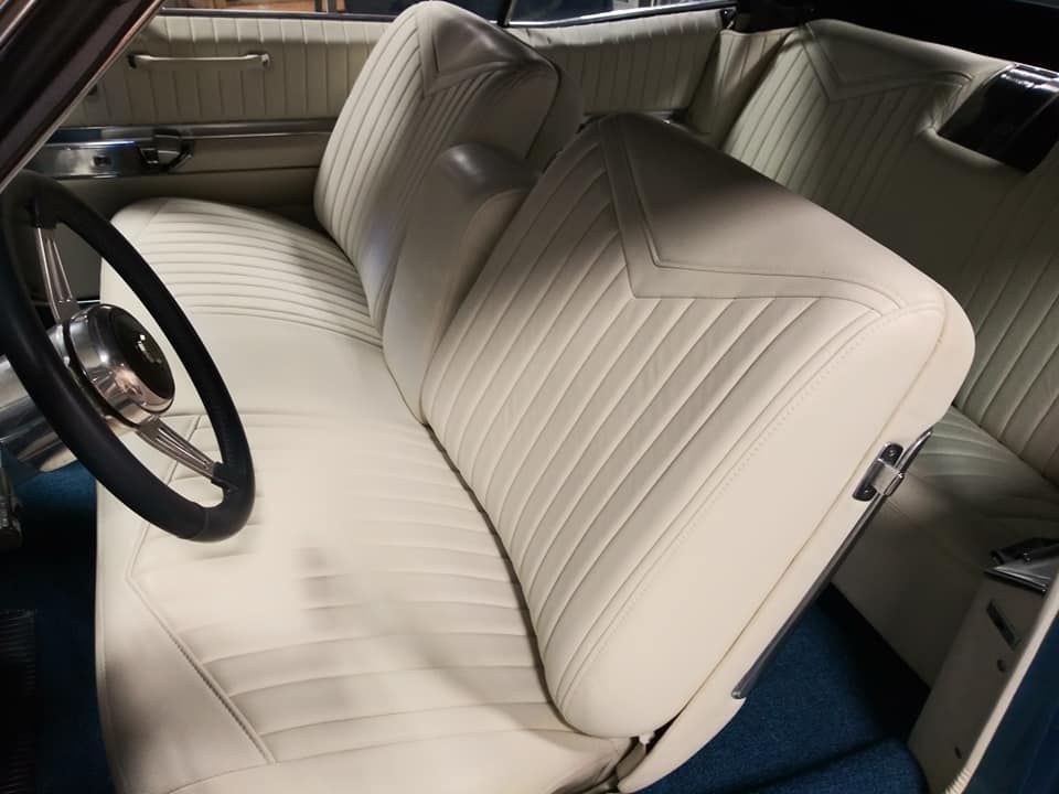 Interior of a vintage car featuring white, vertically-stitched leather bench seats and blue carpet flooring.