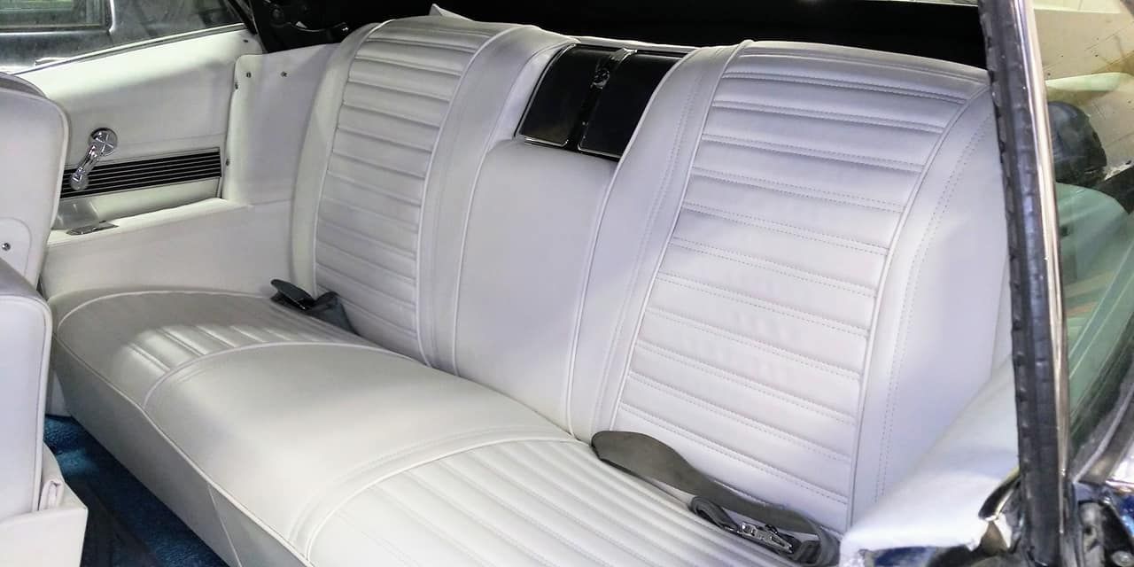 The white vinyl bench seat of a classic car interior with a black center emblem and seat belts.