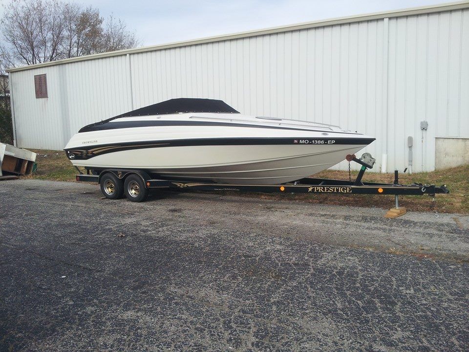 A white and black powerboat with a dark cover sits on a dual-axle trailer in front of a white metal building.