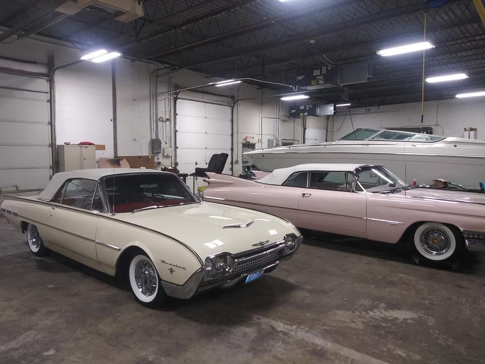 A white Ford Thunderbird and a light pink Cadillac convertible parked inside a large, industrial-style garage.