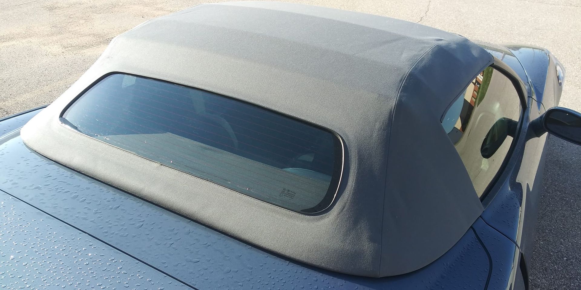 A high-angle view of a dark blue convertible car’s grey fabric roof and rear glass window.